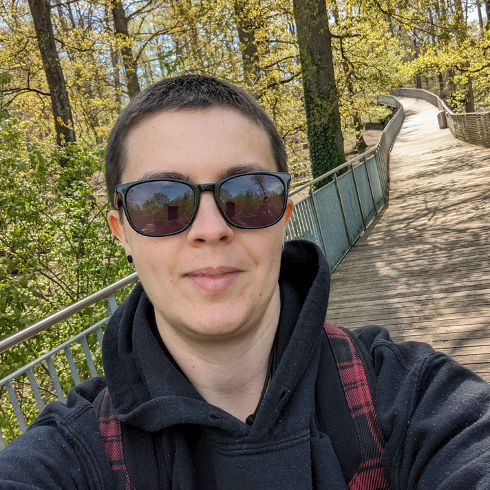 Selfie of a woman with buzz cut black hair and sunglasses on a high wooden walkway in a forest